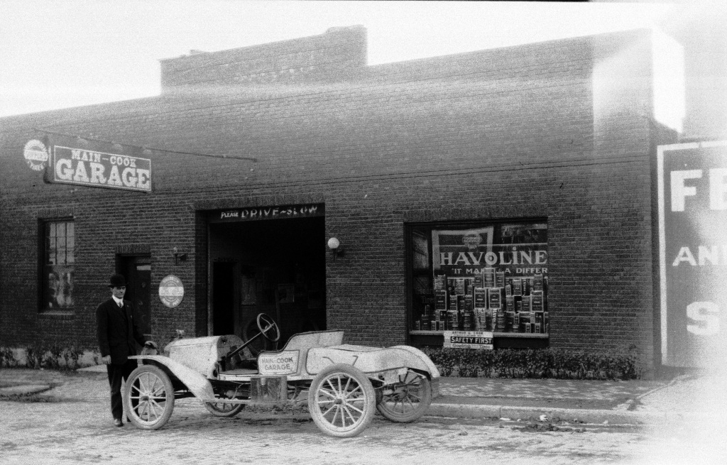Archival photograph of the Main-Cook Garage in Delaware, Ohio. Photo shows the front edifice with an unknown cleanshaven white man in a black suit and bowler hat standing in front with a vehicle from the time period (perhaps 1930s).